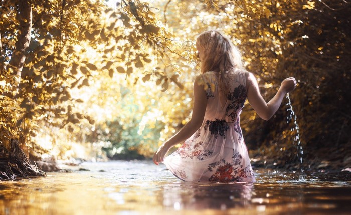 Woman in a dress wading in a stream with sunlight filtering through trees