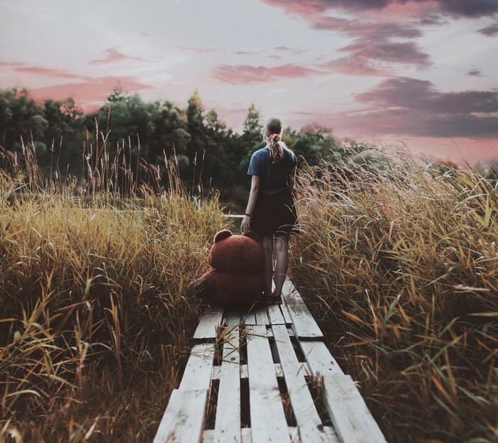 Girl with teddy bear on wooden path in tall grass