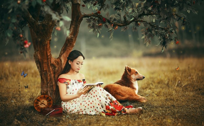 Woman reading a book with a dog under a tree