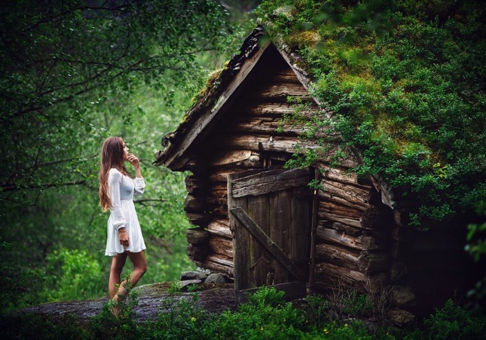 Woman in white dress near a moss-covered wooden hut in a forest