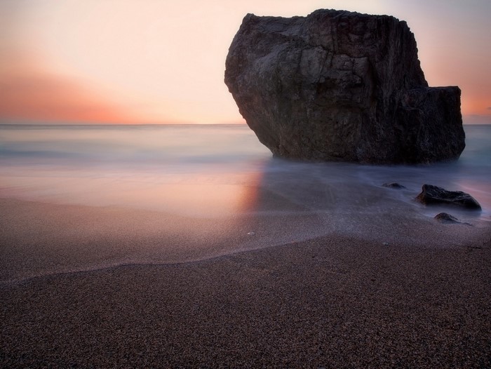 Large rock on a sandy beach at sunset with calm ocean waves