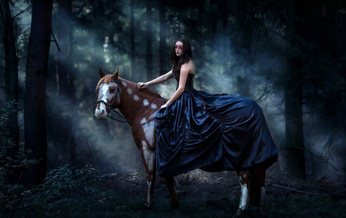 Woman in dark dress riding a spotted horse in a misty forest