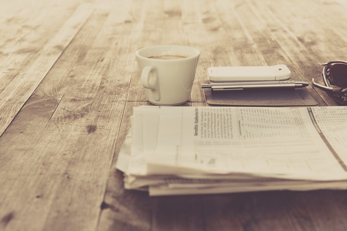 Coffee cup, smartphone, pen, and newspaper on a wooden table