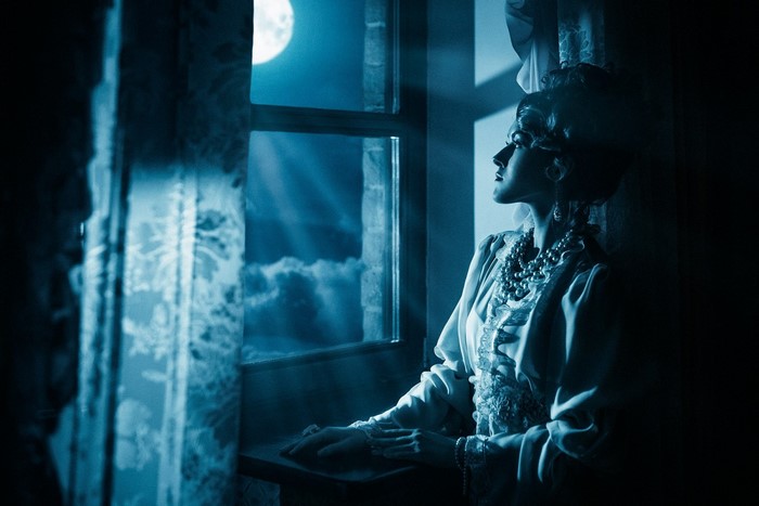 Woman in vintage dress looking out window at moonlight
