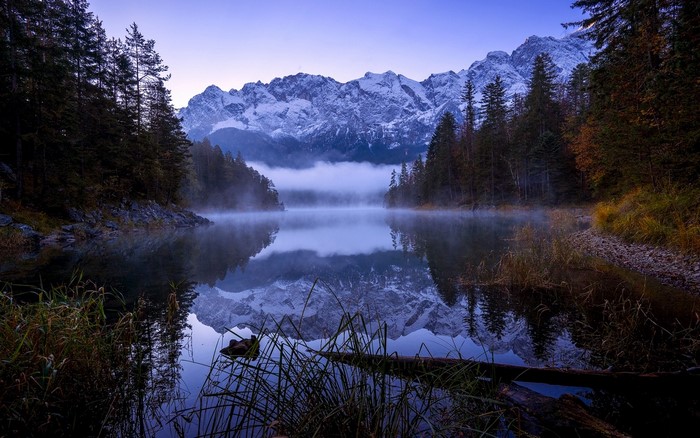 Misty lake reflecting snow-capped mountains and pine forest