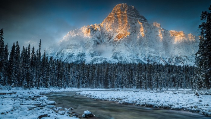 Snowy mountain peak illuminated by morning sunlight over a frozen forest and river