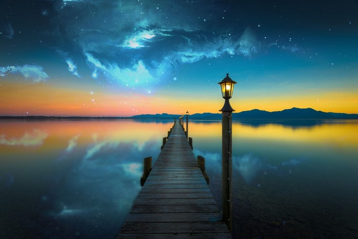 Wooden pier with lampposts over a calm lake at sunset with starry sky