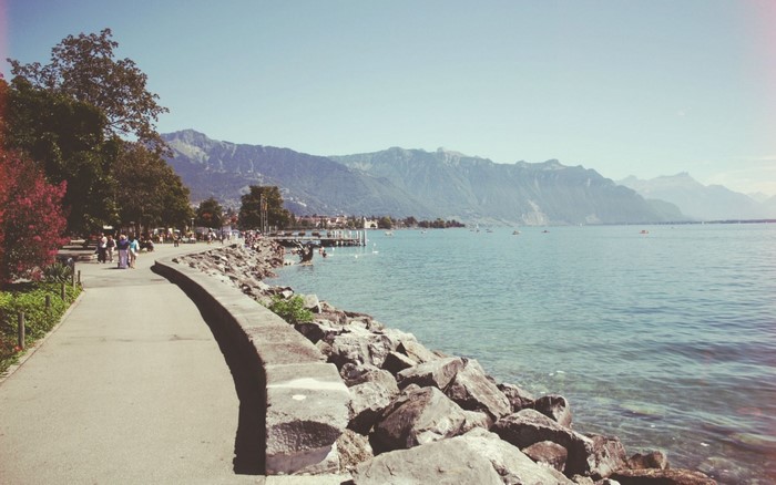 People walking along a paved path by a rocky shore and a lake with mountains in the background