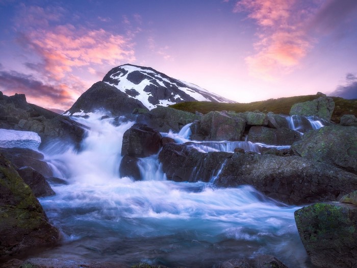 Waterfall flowing over rocks with snow-capped mountain and pink sky