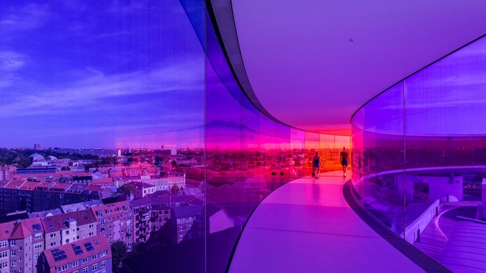 People walking in a colorful glass hallway overlooking a cityscape at dusk