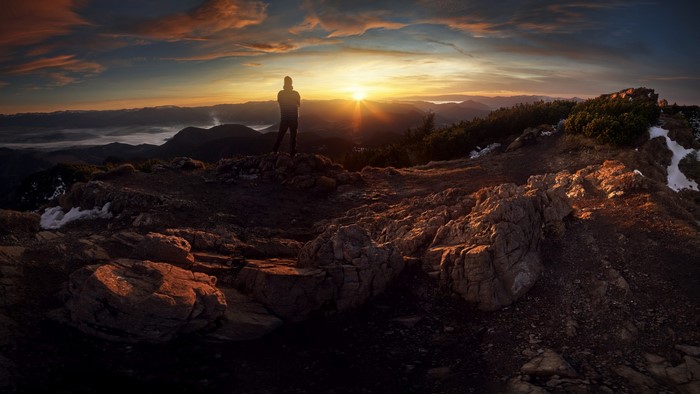 Man on mountain summit watching sunrise over misty landscape
