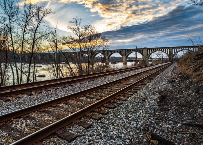 Railroad tracks leading to an arched bridge over a river at sunset