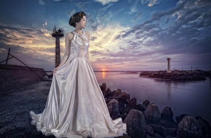 Asian woman in wedding dress by lighthouse at sunset
