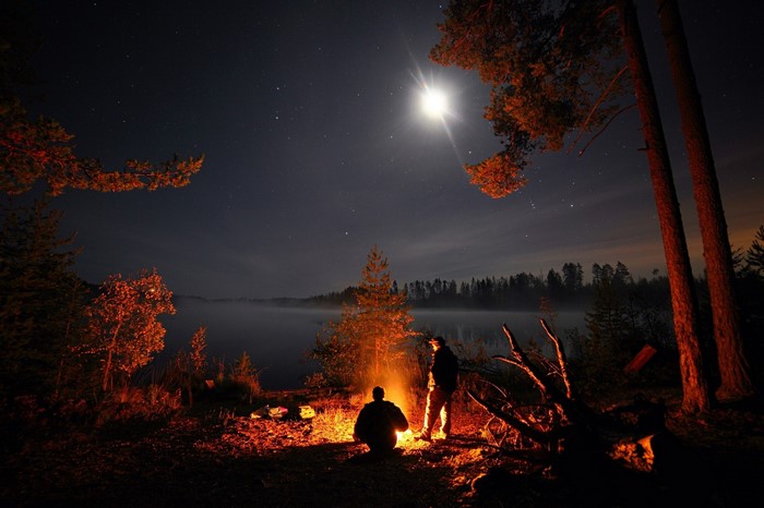 Two people camping by a lake at night with a bonfire
