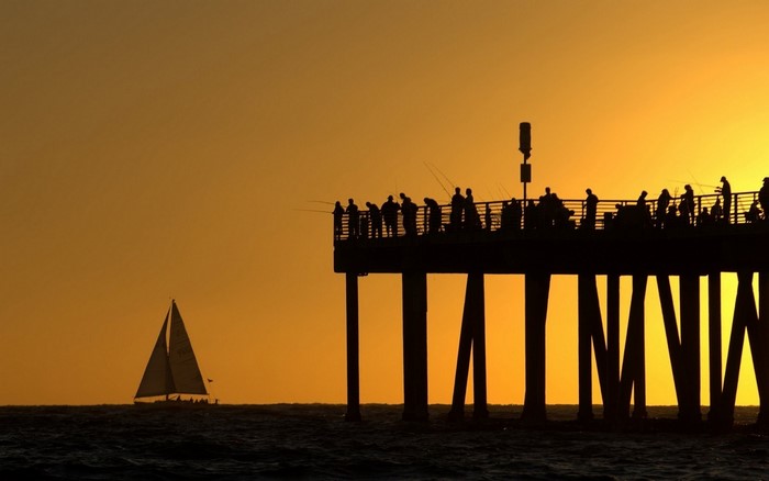 Silhouette of people fishing on a pier at sunset with a sailboat