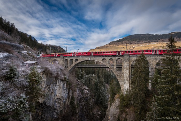 Red train crossing a stone viaduct bridge in a mountainous landscape