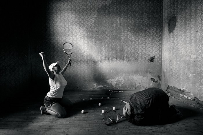 Woman celebrating victory over defeated man in abandoned room with tennis equipment