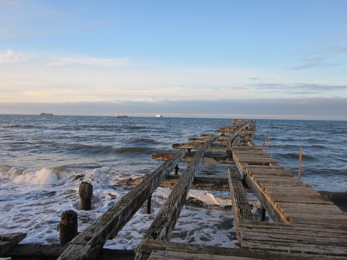 Dilapidated wooden pier extending into the ocean with ships on the horizon