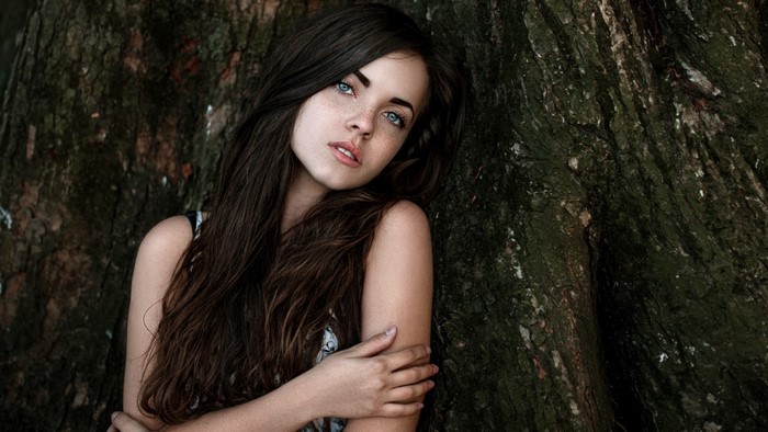 Woman with long brown hair and blue eyes leaning against a tree