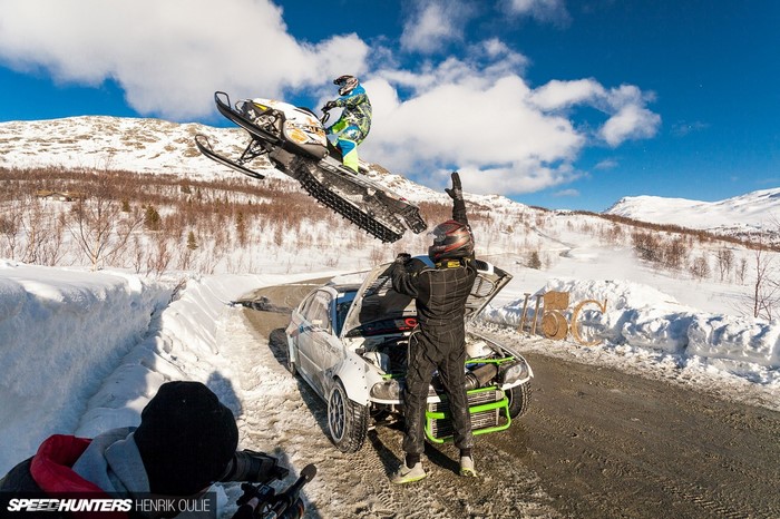 Snowmobile jumps over a car with a person in racing gear
