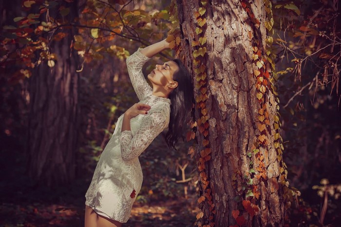 Woman in white lace dress leaning against a tree in autumn forest