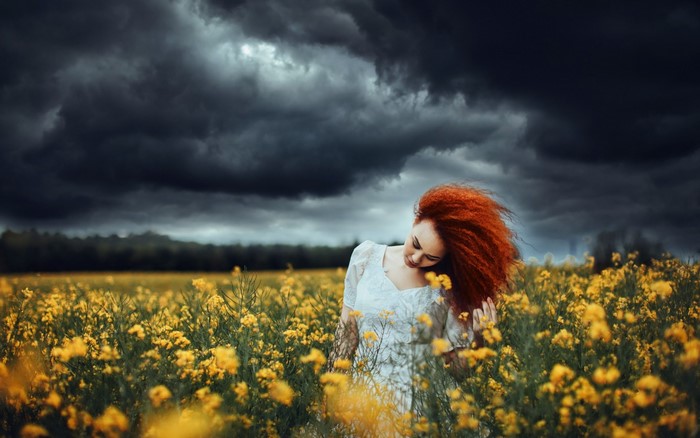 Woman with red hair in a field of yellow flowers under stormy clouds