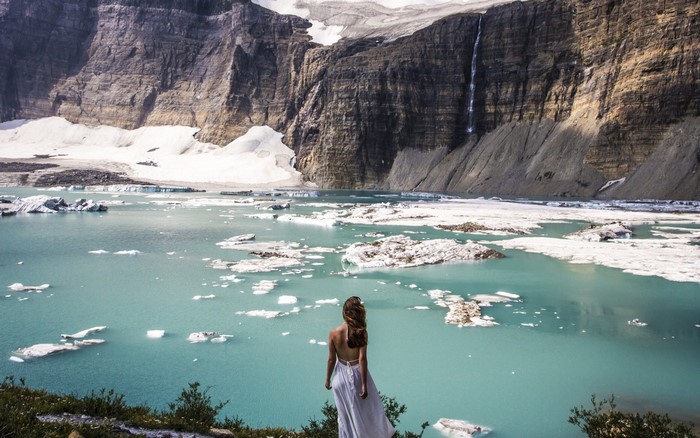 Woman in white dress looking at glacial lake with icebergs and waterfall