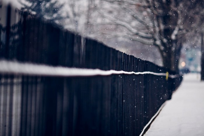 Snow falling on a dark fence along a snowy path