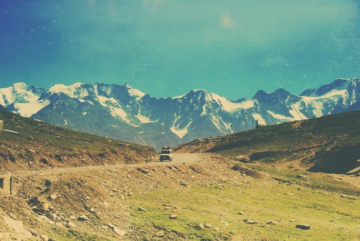 A car drives on a mountain road with snow-capped mountains in the background.