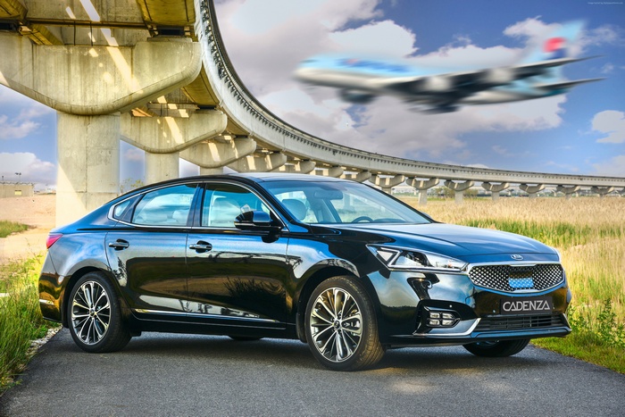Black Kia Cadenza sedan under elevated tracks with airplane
