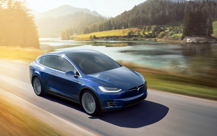 Blue Tesla Model X driving on a road with mountains and a lake