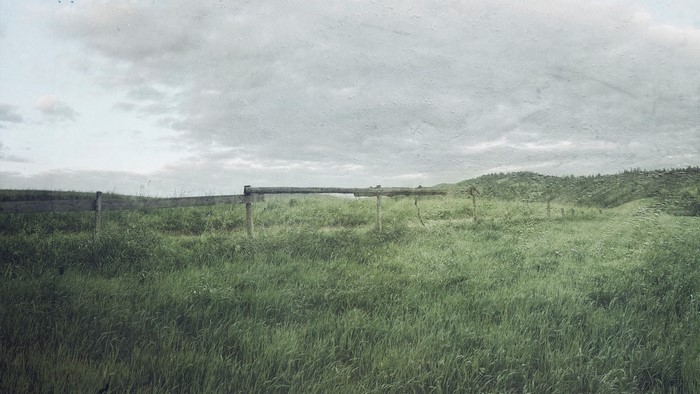 Grassy field with wooden fence and rolling hills under cloudy sky