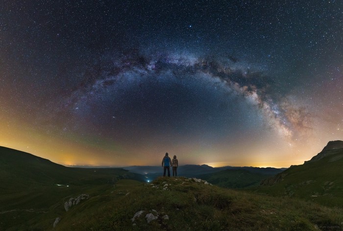 Two people stand on a hill under the Milky Way galaxy