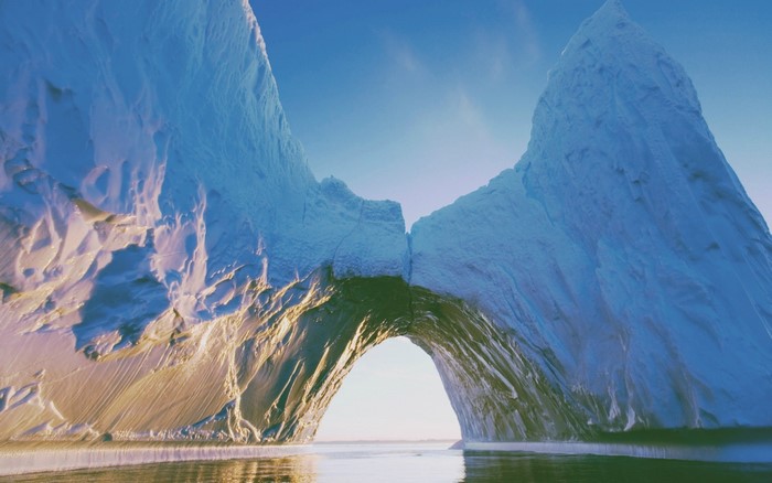 Iceberg arch over calm water under blue sky