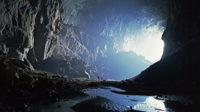 Two people explore a massive cave with sunlight streaming in