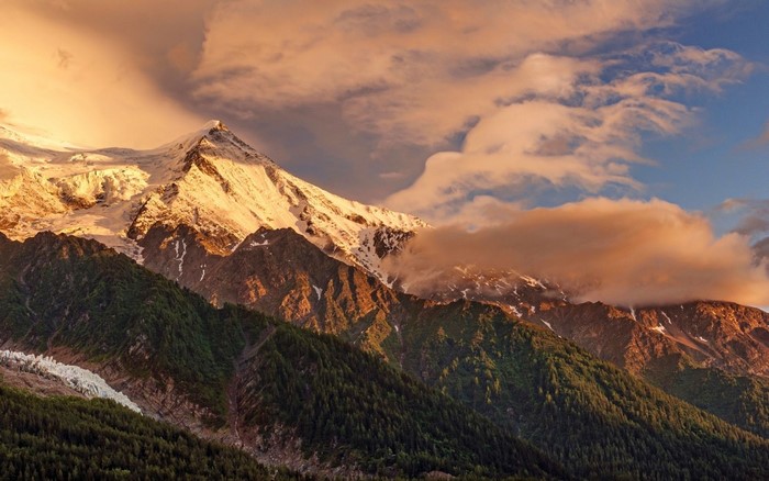 Snowy mountain peak at sunset with clouds and forest