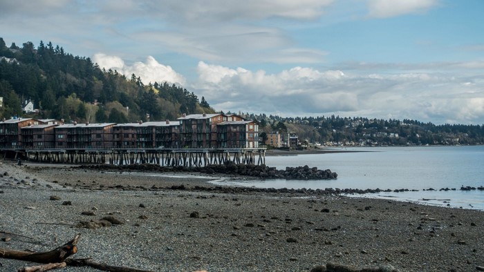 Apartment buildings on stilts over a rocky shore with a bay and forested hills
