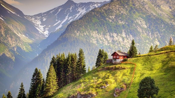 Red house on a grassy mountain slope with snow-capped peaks