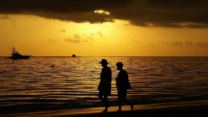 Two people walk on a beach at sunset with a boat offshore.