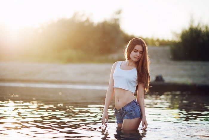 Woman in jean shorts and crop top standing in river at sunset