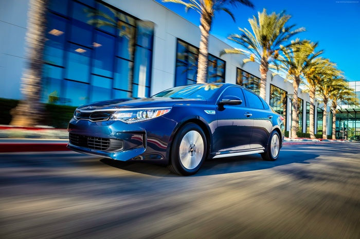 Blue Kia Optima Hybrid driving past modern building with palm trees
