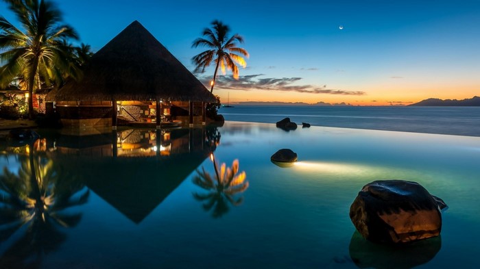 Infinity pool at sunset with thatched bungalow and palm trees
