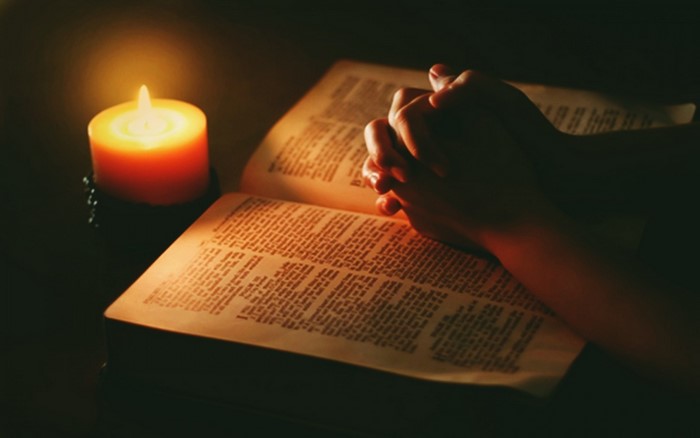 Hands praying over a Bible by candlelight