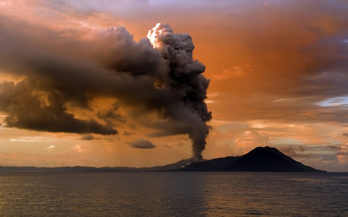 Volcano eruption spewing smoke into the sky over the ocean at sunset