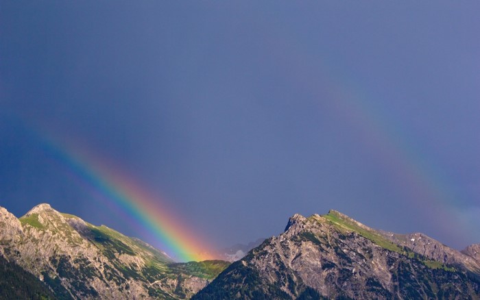 Rainbow over rocky mountains under a dark blue sky
