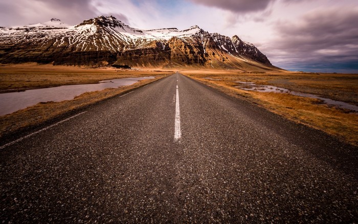 Asphalt road leading to snow-capped mountains under cloudy sky