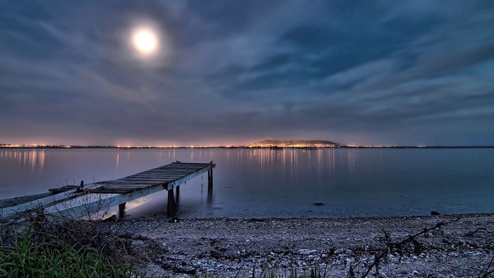Moonlit dock on a calm lake with distant city lights