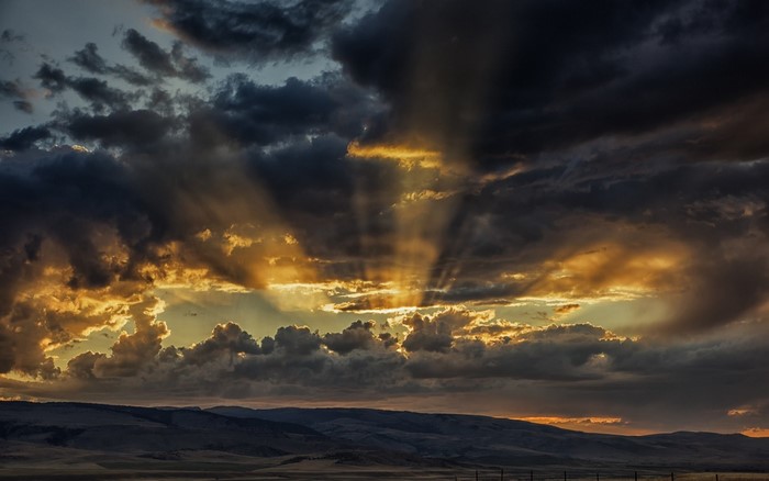 Sun rays piercing through dark clouds over hills at sunset
