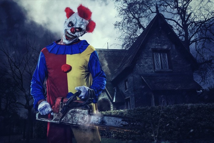 Scary clown with chainsaw in front of a haunted house