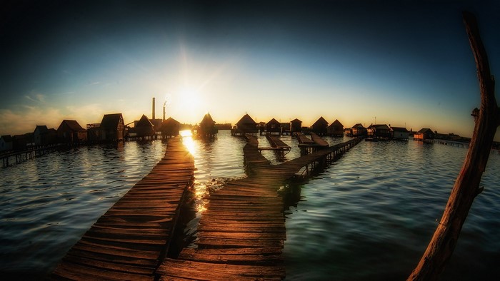 Wooden walkways lead to stilt houses on a lake at sunset